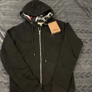 Svart Burberry hoodie med dragkedja - Svart hoodie Burberry med klassiskt rutigt foder i huvan och snygga detaljer som silverfärgad dragkedja och snörning. Tröjan har långa ärmar, ribbade muddar och Burberry-logga på bröstet. Perfekt för dig som vill ha en stilren och lyxig look.