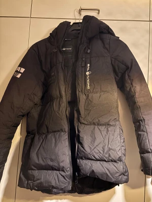 Svart Sail Racing dunjacka S - Svart dunjacka från Sail Racing med huva och coola loggor på bröstet, ärmen och ryggen. Jackan har dragkedja framtill, flera fickor och quiltad design. Perfekt för kalla dagar och har en schysst, sportig look.