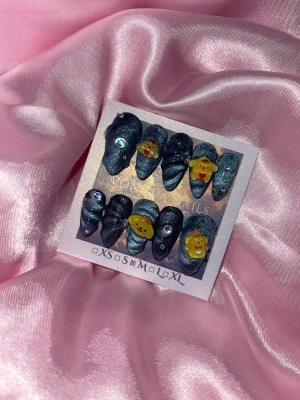 PomGalaxy(M);Medium Almond - Set med press-on naglar i metallic mörkblå färg, dekorerade med små rhinestones och 3D-gula figurer med söta ansikten och hjärtan. Naglarna har en unik form med swirl-mönster och finns i M: 16 mm, 12 mm, 12,5 mm, 11,5 mm, 10 m. Perfekt för dig som vill ha något extra på naglarna.