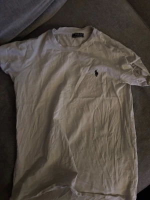 Vit t-shirt från Polo Ralph Lauren - Vit t-shirt från Polo Ralph Lauren med klassisk rund hals och korta ärmar. T-shirten har den ikoniska Polo-loggan broderad i svart på bröstet. Tillverkad i mjuk bomull för skön känsla och enkel stil.