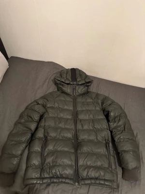  Peak Performance jacka  - Säljer en mörkgrön dunjacka från Peak Performance med huva och svarta detaljer. Jackan har två sidofickor med dragkedja, quiltad design och Peak Performance-logga på bröstet och huvan. Perfekt för kalla dagar och har en clean, sportig look. Storlek 160 Inga repor eller skador på de nästan ny