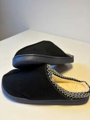 Uggs liknande tofflor - Svarta tofflor med mjuk fuskpäls på insidan och dekorativt silverfärgat mönster runt öppningen. Ovandel i mockaliknande material och platt sula. Perfekta för att hålla fötterna varma hemma under kyliga dagar. Helt nya.