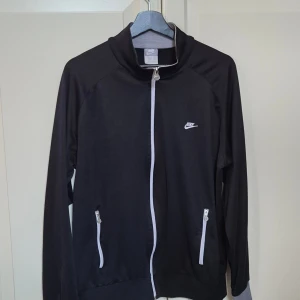 Svart vintage ziptröja från Nike M - Svart vintage ziptröja från Nike med vit dragkedja och vita detaljer vid fickorna. Tröjan har lång ärm, ståkrage och Nike-logga på bröstet. Perfekt för träning eller chill, tillverkad i mjukt syntetmaterial som andas.