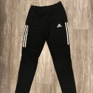 Svarta Adidas träningsbyxor - Svarta träningsbyxor från Adidas med klassiska vita ränder längs sidorna och Adidas-logga på låret. Byxorna har dragkedja vid benslut och är tillverkade i ett syntetmaterial som passar perfekt för sport och träning. Storlek visas i sista bilden