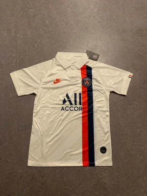 PSG Neymar Jr 10 fotbollströja Nike - Snygg PSG fotbollströja med Neymar Jr och nummer 10 på ryggen. Tröjan är vit med en röd och marinblå rand på framsidan, broderat Nike-märke och PSG-logga. Klassisk krage och korta ärmar. Tillverkad i lätt och ventilerande polyester, perfekt för match eller träning.