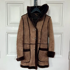 Mocka pälsjacka vintage  - Perfekt skräddad, kort retro päls. Snäv, lite klockad modell med huva. Både söt, stilfull och varm. Helfodrad i fräscht skick. Fuskpäls och mocka som ger ett äkta intryck. Storleken är närmre 34 än 36. Modellen på bilden är strl 36/S. 