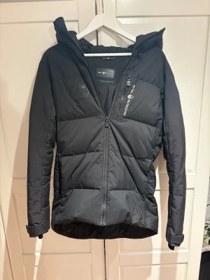 Svart pufferjacka från Sail Racing - Svart pufferjacka från Sail Racing med huva och snygga logodetaljer på bröstet, ärmen och ryggen. Jackan har dragkedja framtill, quiltad design och långärmade ärmar. Perfekt för kalla dagar och har en clean, sportig look. Nypris 3299 mitt pis 1999!