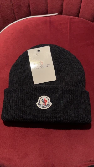 Svart Moncler mössa - Svart ribbstickad mössa från Moncler med klassisk uppvikt kant och broderad logotyp framtill. Tillverkad i mjuk ull som håller dig varm under kalla dagar. Perfekt accessoar för en clean och stilren look.