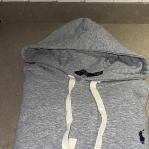 Grå hoodie från Polo Ralph Lauren - Snygg grå hoodie från Polo Ralph Lauren med klassisk broderad logga i marinblått på bröstet. Tröjan har huva med vita snören och ribbade muddar vid ärmslut och nederkant. Nypris: 1799, mitt pris: 849