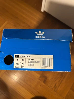 Adidas Everyrn W rosa sneakers - Säljer ett par Adidas Everyrn W sneakers i ljusrosa med vit platt sula och svart detalj på hälen. Skorna har snörning, rund tå och är tillverkade i skinn. Snygg gulddetalj med Adidas-logga på sidan. Perfekt för dig som gillar klassisk streetstil. Strl i lagret: 41 1/3, 43 1/3