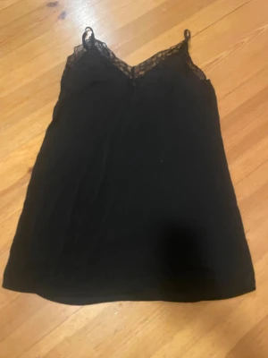 Svart linnetopp med spets - Säljer en svart linnetopp med tunna axelband och snygga spetsdetaljer längs v-ringad hals. Toppen har en enkel och stilren design som är lätt att matcha. Perfekt för dig som gillar en clean look med lite extra edge.