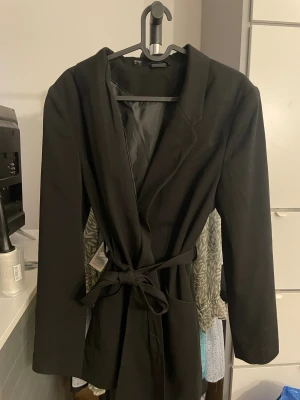 Svart kavaj med bälte - Svart kavaj med klassisk krage och lång ärm, tillverkad i ett slätt tyg med fodrad insida. Jackan har ett matchande bälte i midjan för snygg passform och två stora fickor framtill. Perfekt för dig som gillar stilrena och enkla plagg. Stor i storlek 