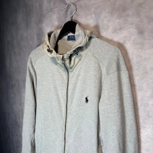 Ljusgrå hoodie från Polo Ralph Lauren - Snygg ljusgrå hoodie från Polo Ralph Lauren med dragkedja och huva med snörning. Tröjan har en klassisk passform och är tillverkad i mjuk bomull. Ikoniska loggan broderad på bröstet. Perfekt för chill dagar eller när du vill vara casual.