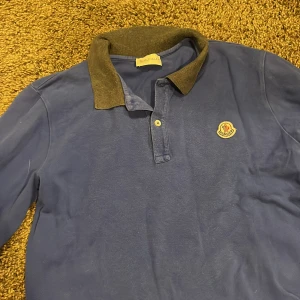 Blå långärmad pikétröja från Moncler - Snygg blå långärmad pikétröja från Moncler med kontrastfärgad krage och muddar i mörkgrått. Tröjan har klassisk pikéknäppning med två knappar och Moncler-logga broderad på bröstet. Perfekt för dig som gillar stilren och sportig look.