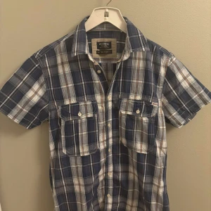 Blårutig kortärmad skjorta Jack & Jones - Säljer en blårutig kortärmad skjorta från Jack & Jones Vintage. Skjortan har två bröstfickor med lock och knappar, klassisk krage och är gjord i mjuk bomull. Perfekt för dig som gillar avslappnad stil med lite edge. Vid frågor hör av dig, Mvh Oscar
