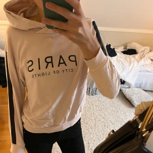 Rosa/Beige hoodie med svart tryck och ränder - Säljer en super skön beige hoodie med svart text 'PARIS CITY OF LIGHTS' på bröstet och svarta ränder längs ärmarna. Tröjan har huva, ribbade muddar och är långärmad. Perfekt för en chill och snygg look. Hoodien är lite nopprig (se bild 3)