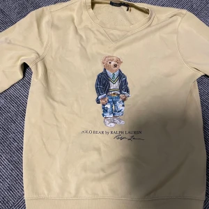 Beige Polo Bear tröja från Ralph Lauren - Snygg beige tröja från Ralph Lauren med ikoniska Polo Bear-trycket på bröstet. Tröjan har rund halsringning och långa ärmar. Mjuk insida och klassisk passform, perfekt för dig som gillar streetstyle och preppy vibes.