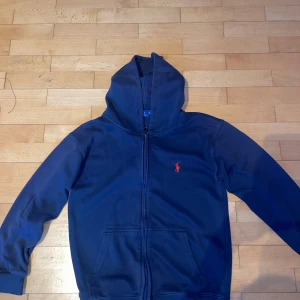 Marinblå hoodie från Polo Ralph Lauren - Snygg marinblå hoodie från Polo Ralph Lauren med dragkedja och klassisk röd broderad logga på bröstet. Tröjan har huva, känguruficka och är tillverkad i mjuk bomull. Perfekt för en chill och stilren look.