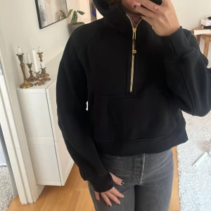 Lululemon hoodie - Svart hoodie från Lululemon med guldig half zip-dragkedja och stor känguruficka framtill. Tröjan har huva med Lululemon-logga. 