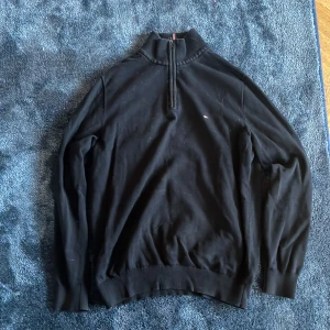 Svart half zip tröja Tommy Hilfiger - Svart långärmad tröja från Tommy Hilfiger med half zip och ribbad krage. Diskret logga på bröstet och snygg detalj med röd-vit rand på insidan av kragen. Perfekt för dig som gillar stilrena och klassiska plagg.