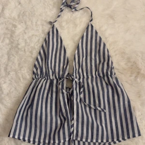 Randig blåvit halterneck linnetopp - Säljer en snygg randig halterneck topp i blått och vitt. Toppen har knytning i nacken och vid bysten, samt en luftig passform nedtill. Perfekt för varma dagar och ger en chill sommarvibe. Materialet känns lätt och svalt mot huden.