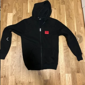 Svart hoodie från Hugo Boss med dragkedja - Svart hoodie från Hugo Boss med röd logotyp på bröstet. Tröjan har huva, hel dragkedja framtill och känguruficka. Tillverkad i mjukt material, perfekt för en avslappnad stil.