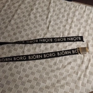 Svart Björn Borg bälte med logga - Svart bälte från Björn Borg med tydlig vit logotyp längs hela bandet. Bältet har ett stilrent metallspänne i silverfärg där märket också är ingraverat. Tillverkat i slitstarkt syntetmaterial