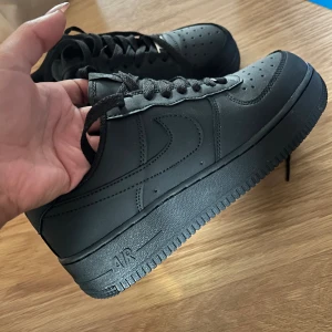 Svarta Nike Air Force 1 sneakers - Säljer ett par helsvarta Nike Air Force 1 sneakers med klassisk design. Skorna har snörning, rund tå och är tillverkade i skinn med en platt sula. Perfekta för dig som gillar en clean och tidlös look. Säljes pågrund av fel beställning av storlek.