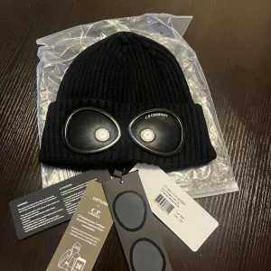 Svart Goggle-mössa från C.P. Company - Svart ribbstickad mössa från C.P. Company med ikoniska goggle-linser framtill. Mössan är tillverkad i mjuk ull och har en uppvikt kant. Perfekt för dig som vill sticka ut med en streetwear-inspirerad look.