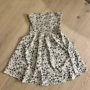 Blommig tubklänning i bomull - Supersöt kort blommig tubklänning i vit bomull med smock upptill. Klänningen har ett härligt mönster med små blommor och är perfekt för varma dagar. Den är ärmlös och har en luftig passform som känns både bekväm och trendig.