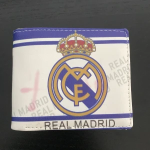 Real Madrid plånbok i vitt och blått - Plånbok med Real Madrid-logga och text i blått och guld på vit bakgrund. Flera kortfack och fönster för ID. Snygg sportig design för dig som älskar fotboll och vill visa ditt lag. Tillverkad i syntetmaterial.