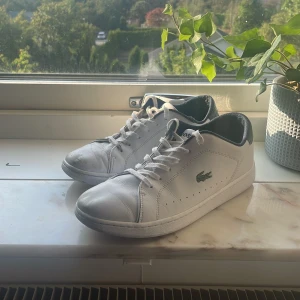 Vita sneakers från Lacoste i skinn - Snygga vita sneakers från Lacoste i skinn med klassisk grön krokodil-logga på sidan. Skorna har snörning, rund tå och vit platt sula. Insidan är fodrad i mörkblått tyg och Lacoste-logga syns på både häl och plös. Perfekta för en clean och sportig look.