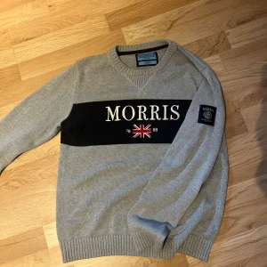 Grå stickad tröja från Morris Co - Snygg grå stickad tröja från Morris Co i storlek medium. Tröjan har ett svart band över bröstet med broderad logga och brittisk flagga, samt patch på ärmen. Ribbstickade muddar vid hals, ärmslut och nederkant. Perfekt för dig som gillar klassisk stil.