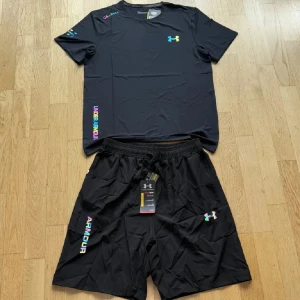 Svart t-shirt från Under Armour - Svart t-shirt från Under Armour med färgglada loggor och tryckdetaljer på bröstet, ärmen och sidan. T-shirten är kortärmad och har en sportig vibe, perfekt för träning. Materialet är lätt och andas, vilket gör den skön att ha på sig under aktiva dagar.
