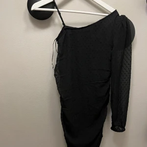 Svart oneshoulder kortklänning - Snygg svart kortklänning med ett axelband och en lång ärm. Klänningen har diskret prickigt mönster i tyget och är figurnära med lätt rynkade detaljer längs sidorna. Perfekt för dig som vill sticka ut med en unik och trendig look.