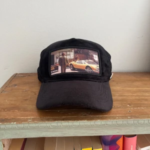 Svart Maggiore keps C2 - Svart keps från Maggiore med mesh på sidorna och bak, justerbart spänne i skinnimitation och coolt motiv med bil och person framtill. Märkets logga syns både invändigt och som metallmärke på sidan. Perfekt för dig som gillar bilar och streetstyle.