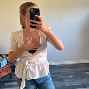 Vit omlottblus med puffärm - Säljer en vit omlottblus med korta puffärmar och knyt i midjan. Blusen har v-ringad hals och ett lätt rynkat parti nedtill som ger en söt siluett. Perfekt till jeans eller kjol för en fräsch look. Otrolig snygg men för liten för mig över bysten som har D i kupa