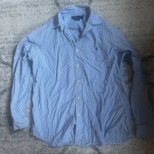 Blåvit randig skjorta från Ralph Lauren - Säljer en klassisk blåvit randig skjorta från Ralph Lauren med broderad logga på bröstet. Skjortan är i 100% bomull med stretch och har långa ärmar samt knappar framtill. Perfekt för dig som gillar stilrena och tidlösa plagg.