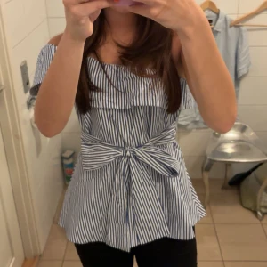 Randig offshoulder blus med rosett - Säljer en blåvit randig blus med offshoulder och stor rosett framtill. Toppen har en volang upptill och är figursydd i midjan för en snygg siluett. Perfekt för dig som gillar trendiga detaljer och somrig stil.