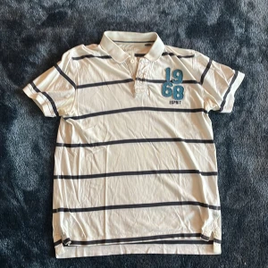 Randig pikétröja från Esprit - Vit pikétröja från Esprit med mörkblå horisontella ränder och broderad detalj med siffrorna 1968 på bröstet. Klassisk krage med knappar och mjukt bomullsmaterial. Perfekt för en avslappnad och stilren look.