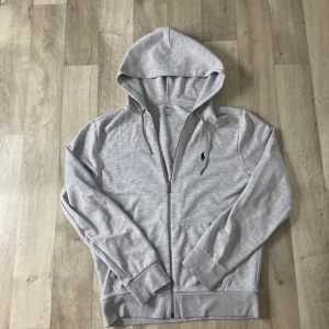 Grå hoodie från Polo Ralph Lauren - Snygga ljusgrå hoodie från Polo Ralph Lauren med dragkedja och klassisk huva. Tröjan har långa ärmar, ribbade muddar och en broderad marinblå logga på bröstet. Perfekt för chill dagar och enkel att matcha med jeans eller joggers. Skicket är 10/10 och använd FÅTAL GÅNGER!