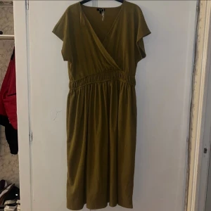 Olivgrön omlottklänning från C&A - Olivgrön omlottklänning från C&A med korta ärmar och resår i midjan. Klänningen har v-ringning och ett mjukt, lätt tyg som ger en avslappnad känsla. Perfekt för dig som gillar enkel men snygg stil.