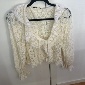 Vit spetsblus från Gina Tricot - Romantisk vit blus i spets från Gina Tricot med långa ärmar och v-ringning. Blusen har volangdetaljer längs kragen och framtill samt knytband vid halsen. Den är genombruten och har en feminin, bohemisk känsla.