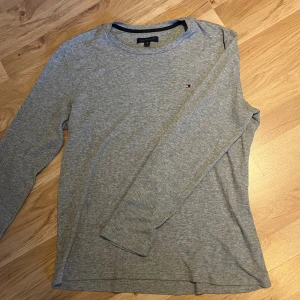 Grå långärmad tröja Tommy Hilfiger - Säljer en stilren grå långärmad tröja från Tommy Hilfiger. Tröjan har ett diskret broderat logomärke på bröstet och är tillverkad i ett mjukt bomullsmaterial med lätt ribbad struktur. Perfekt för en clean och avslappnad look.