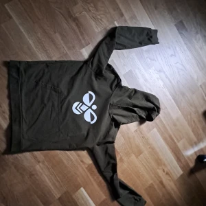 Olivgrön hoodie från Hummel - Säljer en olivgrön hoodie från Hummel med stor vit logga på bröstet. Tröjan har huva, lång ärm och en klassisk känguruficka framtill. Tillverkad i mjuk bomull som är skön att ha på sig. Perfekt för dig som gillar sportig och avslappnad stil.
