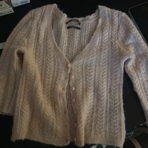 Ljusrosa kofta med knappar från Gina Tricot - Supermysig ljusrosa kofta från Gina Tricot med stickat mönster och v-ringning. Koftan har fina rosa knappar framtill och är gjord i ett fluffigt, stickat material. Perfekt att slänga på sig när det är lite kyligt ute.