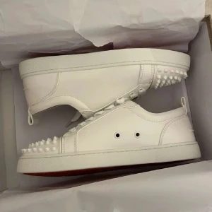 Vita Louboutin sneakers med nitar - Säljer ett par vita sneakers från Christian Louboutin i skinn med ikoniska röda sulor och coola nitar på tån och hälen. Skorna har snörning och en stilren, modern design som sticker ut. Perfekta för dig som vill ha något extra till din outfit.