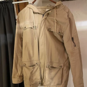 Beige trenchcoat från Resteröds M - Snygg beige trenchcoat från Resteröds i storlek M. Jackan har huva med snörning, fyra stora fickor med knappar framtill och dragkedja. Ärmarna är långa och det finns en extra ficka med dragkedja på ena ärmen. Perfekt för dig som gillar stilren och funktionell design. Priset är ej hugget i sten.