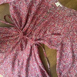 Blommig röd blus - Supersöt blommig blus i rött, vitt och rosa från 365 Sun Shine. Tröjan är sval, och passar till allt! Midjan markeras med ett knytband för extra snygg passform.