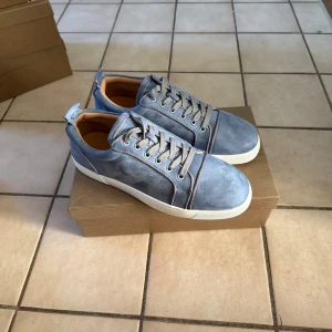 Blå mocka sneakers från Louboutin storlek 42 - Snygga sneakers i ljusblå mocka från Louboutin med klassisk röd sula och vita kanter. Skorna har rund tå, snörning och diskreta detaljer i silver. Insidan är beige och materialet känns lyxigt och mjukt. Perfekta för dig som vill sticka ut med stil.